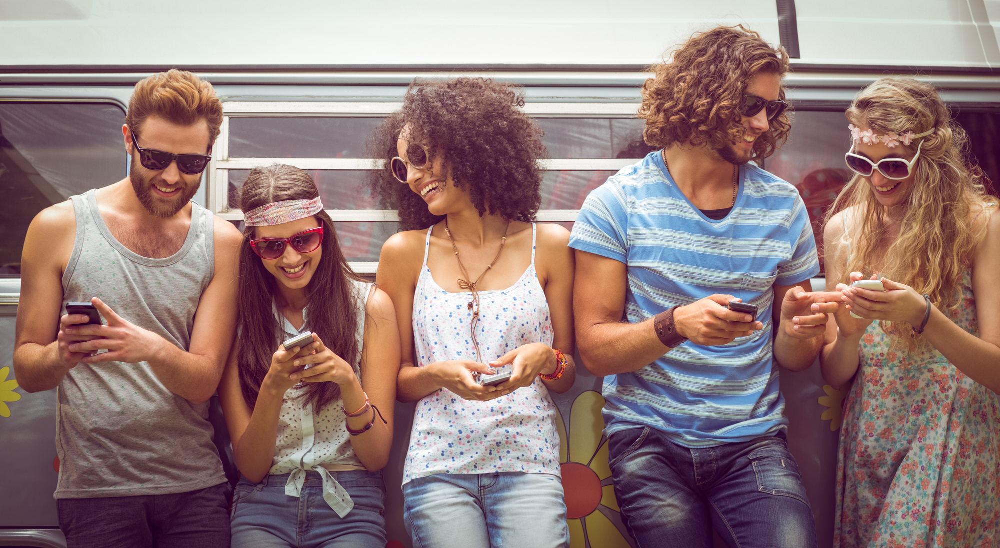 five young friends in summer attire leaning on the side of a van, smiling and looking at their smartphones